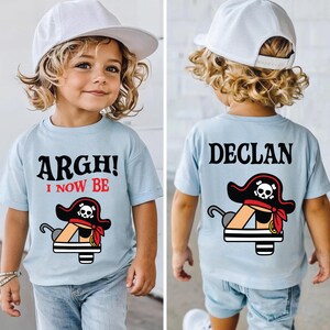 Op de afbeelding: Lichtblauw T-shirt met een piratenthema. De voorkant toont de tekst "ARGH! I NOW BE" boven een piratenhoed en haak illustratie. De achterkant van het shirt toont de naam "DECLAN" en dezelfde piratengrafiek.
