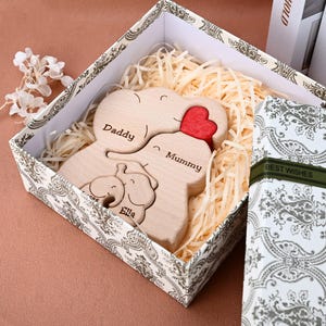 May include: A wooden family elephant puzzle with the words "Daddy", "Mummy", and "Ella" engraved on it. The puzzle includes a red heart and is nestled in a decorative box with "Best Wishes" on the side.