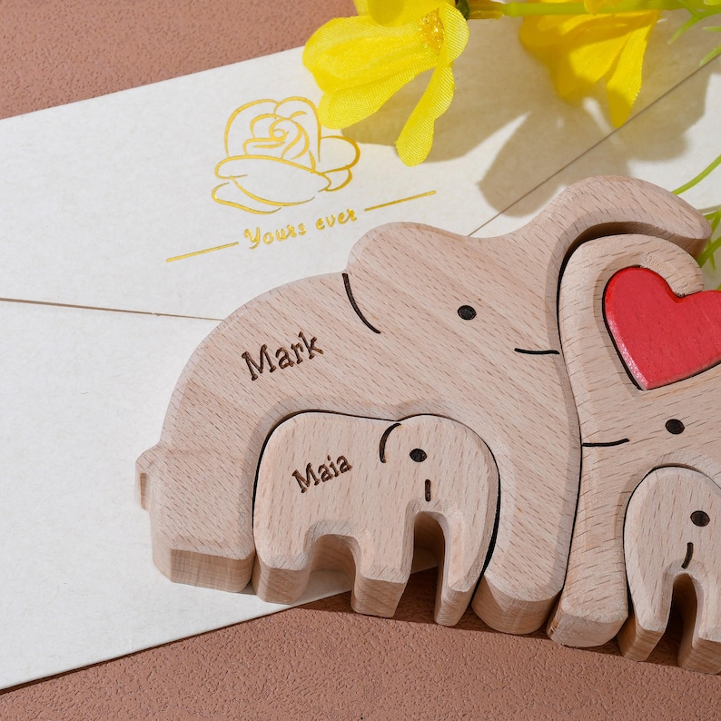 May include: Wooden elephant family puzzle with the names "Mark" and "Maia" engraved. The largest elephant has a red heart. A card with a gold rose and the words "Yours ever" is in the background.