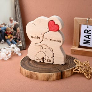 May include: A wooden family puzzle in the shape of elephants. The puzzle features the words "Daddy", "Mummy", "Lucas", and "Ella". A red heart is at the top. The puzzle is displayed on a wooden slice.