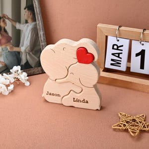 May include: A wooden puzzle featuring two elephants embracing, with a red heart accent. The names "Jason" and "Linda" are engraved on the puzzle. A wooden calendar displays "MAR 1". A framed photo and a star-shaped decoration are also visible.