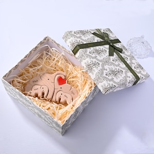 May include: A wooden elephant family set inside a decorative gift box. The elephants are engraved with names and a red heart. The box is patterned with a green ribbon and bow.