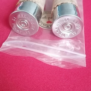 May include: Two bottle stoppers featuring natural cork tops attached to silver-coloured metal bases. The bases are repurposed shotgun shell casings with the text Fiocchi 12 Italy stamped on the silver metal ends. They are inside a clear plastic bag.