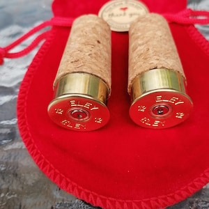 May include: Two gold shotgun shell bottle stoppers with cork tops. The stoppers are on a red velvet bag with a white tag that says "Thank You".