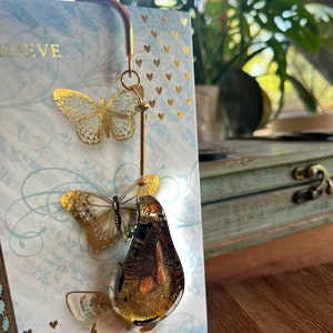 May include: Gold-tone bookmark with a pear-shaped glass charm and two butterfly accents. The charm has a gold leaf design and the butterflies are translucent with gold accents. The bookmark is attached to a card with the words "Believe" and "Hope" printed on it.