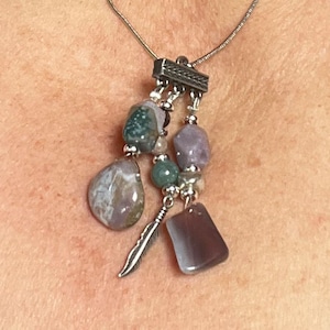May include: A silver chain necklace with a silver pendant featuring a cluster of purple, green, and white stones. The pendant is attached to the chain with a silver bar.