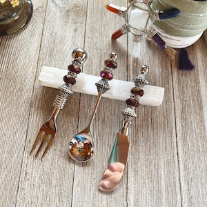 Könnte beinhalten: Drei silberne Besteckteile mit verzierten, perlenbesetzten Griffen. Die Griffe sind aus Silber und haben dunkelviolette Perlen. Die Besteckteile sind eine Gabel, ein Löffel und ein Buttermesser. Die Besteckteile liegen auf einer weißen, rechteckigen Oberfläche.