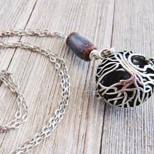 May include: A silver chain necklace with a tree of life pendant and a colorful clay bead. The pendant is a hollow sphere with a black lava rock inside.