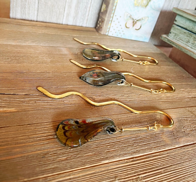 May include: Four gold bookmark holders with glass accents. The glass accents are different colors and patterns. The bookmark holders are on a wooden surface.