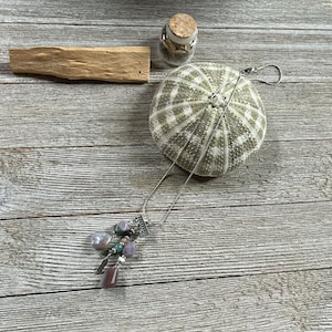 May include: A silver chain necklace with a pendant featuring multiple purple, green, and brown stones. The necklace is laying on a wooden surface with a sea urchin and a small glass jar with a cork lid.