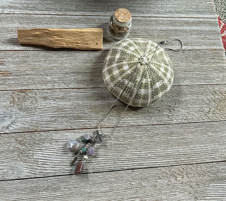 May include: A silver chain necklace with a pendant featuring multiple purple, green, and brown stones. The necklace is laying on a wooden surface with a sea urchin and a small glass jar with a cork lid.