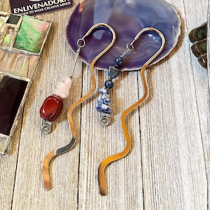 May include: Two handcrafted metal bookmarks with decorative stone beads. One bookmark features red and pink stones, while the other has blue and white stones. The bookmarks have a wavy design and small silver charms at the bottom.