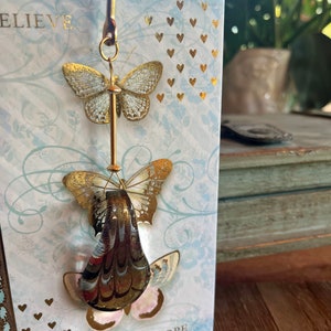 May include: A gold metal bookmark with two gold butterflies hanging from it. The butterflies are on a white background with gold hearts and the words "Believe" and "Hope".