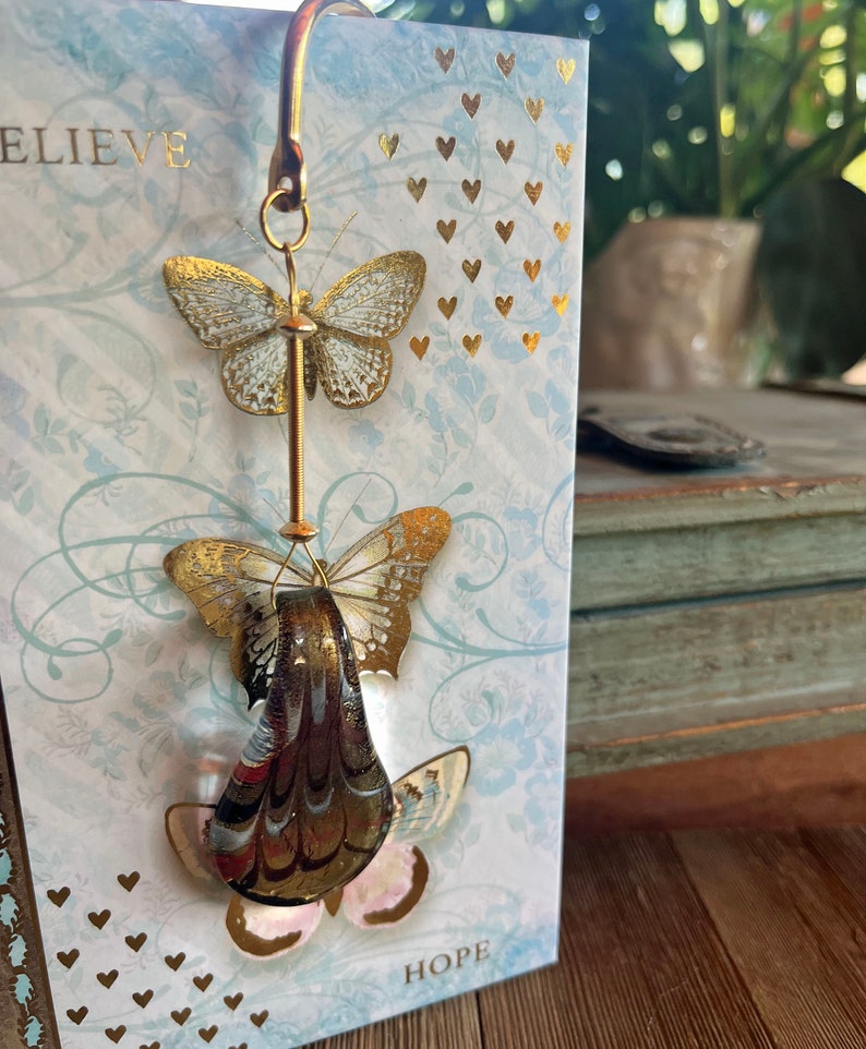 May include: A gold metal bookmark with two gold butterflies hanging from it. The butterflies are on a white background with gold hearts and the words "Believe" and "Hope".