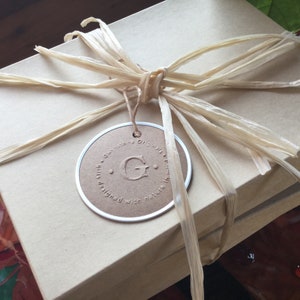 K&ouml;nnte beinhalten: Eine braune Geschenkbox mit einem beigefarbenen Band und einem runden Etikett, auf dem "Gwinstone Originals" mit einem gro&szlig;en "G" in der Mitte steht. Das Etikett sagt auch "silk designed with nature in mind."