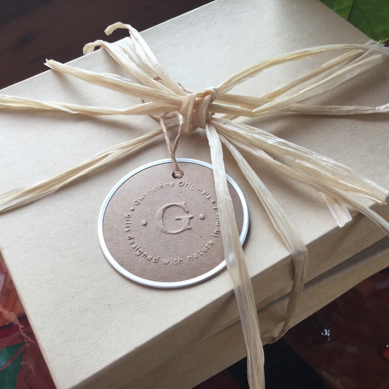 K&ouml;nnte beinhalten: Eine braune Geschenkbox mit einem beigefarbenen Band und einem runden Etikett, auf dem "Gwinstone Originals" mit einem gro&szlig;en "G" in der Mitte steht. Das Etikett sagt auch "silk designed with nature in mind."