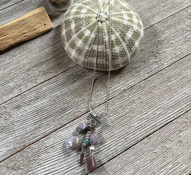 May include: A silver chain necklace with a multi-colored stone pendant. The pendant features a variety of stones, including pink, green, and brown. The necklace is displayed on a wooden surface.