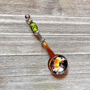 Könnte beinhalten: Ein dekorativer Silberlöffel mit einer runden Schüssel und einem langen Griff. Der Griff hat eine grün-gelbe Glasperle und eine silberne Kugel an der Spitze. Der Löffel ist zum Servieren oder Ausstellen bestimmt.