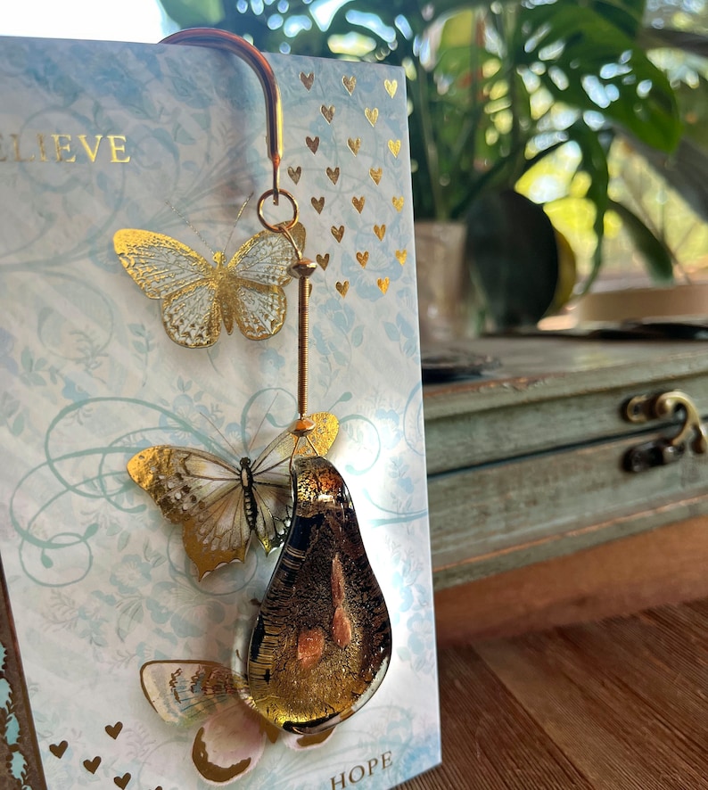 May include: Gold-tone bookmark with a pear-shaped glass charm and two butterfly accents. The charm has a gold leaf design and the butterflies are translucent with gold accents. The bookmark is attached to a card with the words "Believe" and "Hope" printed on it.