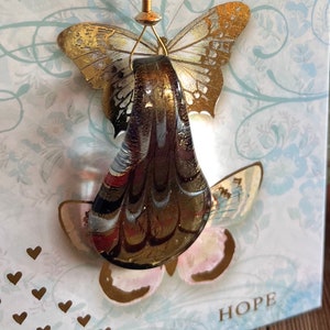 May include: A glass ornament shaped like a teardrop with a gold and brown marbled design. The ornament is hanging from a gold wire and is decorated with two gold butterflies. The word "HOPE" is printed on the card below the ornament.