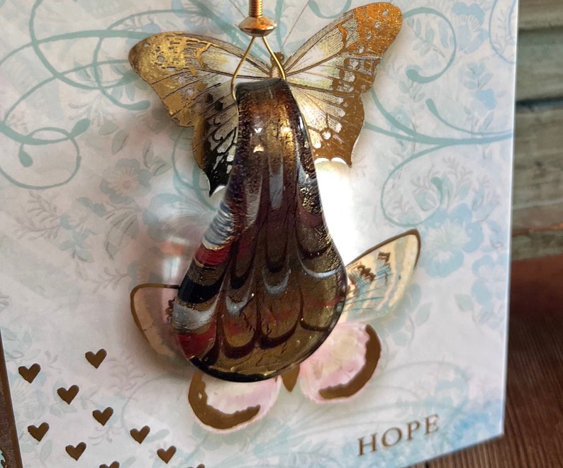 May include: A glass ornament shaped like a teardrop with a gold and brown marbled design. The ornament is hanging from a gold wire and is decorated with two gold butterflies. The word "HOPE" is printed on the card below the ornament.