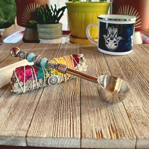 May include: A silver spoon with a beaded handle and a lion charm rests on a white sage smudge stick with colorful flower petals. The spoon is on a wooden surface.