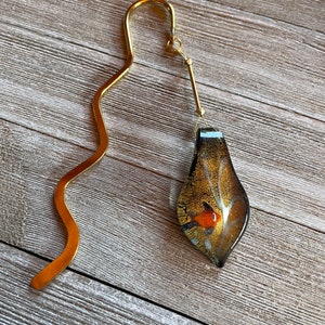May include: A gold bookmark with a wavy design and a glass pendant. The pendant is a teardrop shape with a brown, orange, and white design.
