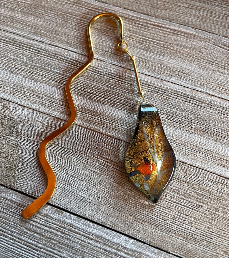 May include: A gold bookmark with a wavy design and a glass pendant. The pendant is a teardrop shape with a brown, orange, and white design.