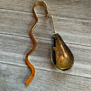 May include: A gold bookmark with a wavy metal design and a teardrop-shaped glass charm with gold flecks.