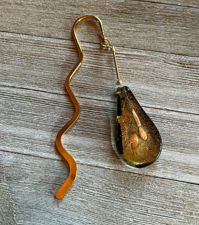 May include: A gold bookmark with a wavy metal design and a teardrop-shaped glass charm with gold flecks.
