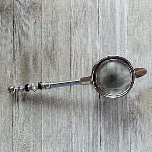 May include: A stainless steel tea infuser with a mesh basket and a long handle. The handle features decorative black beads and silver accents. The infuser is designed for steeping loose leaf tea.