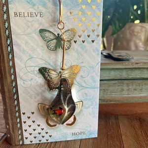 May include: A gold bookmark with three glass butterflies. The butterflies are layered on top of each other and have gold and black accents. The bookmark is hanging from a book with the words "Believe" and "Hope" on the cover.