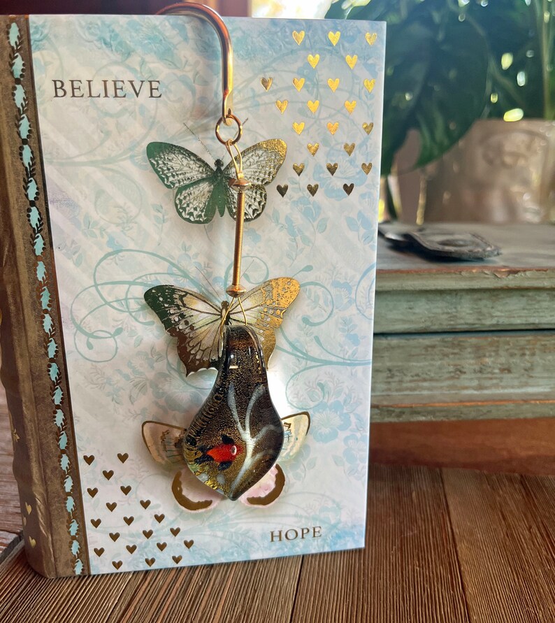 May include: A gold bookmark with three glass butterflies. The butterflies are layered on top of each other and have gold and black accents. The bookmark is hanging from a book with the words "Believe" and "Hope" on the cover.