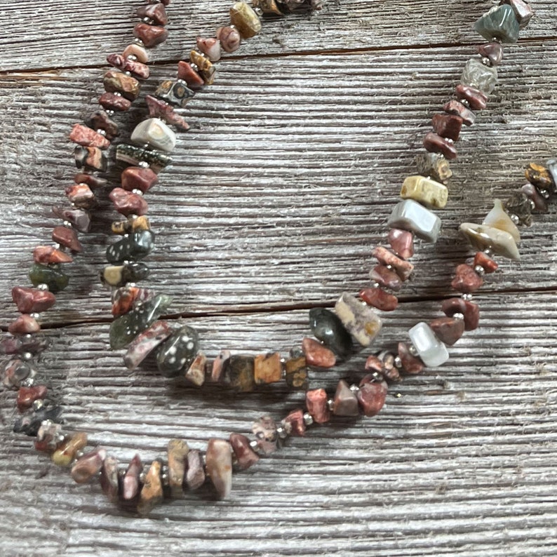 May include: A multi-strand necklace made of various shades of brown, red, and green stones. The stones are irregularly shaped and have a natural, polished finish. The necklace is strung on a silver wire.