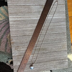 May include: A silver chain necklace with a round, gray and white agate pendant. The necklace is laying on a wooden surface with a ruler next to it.