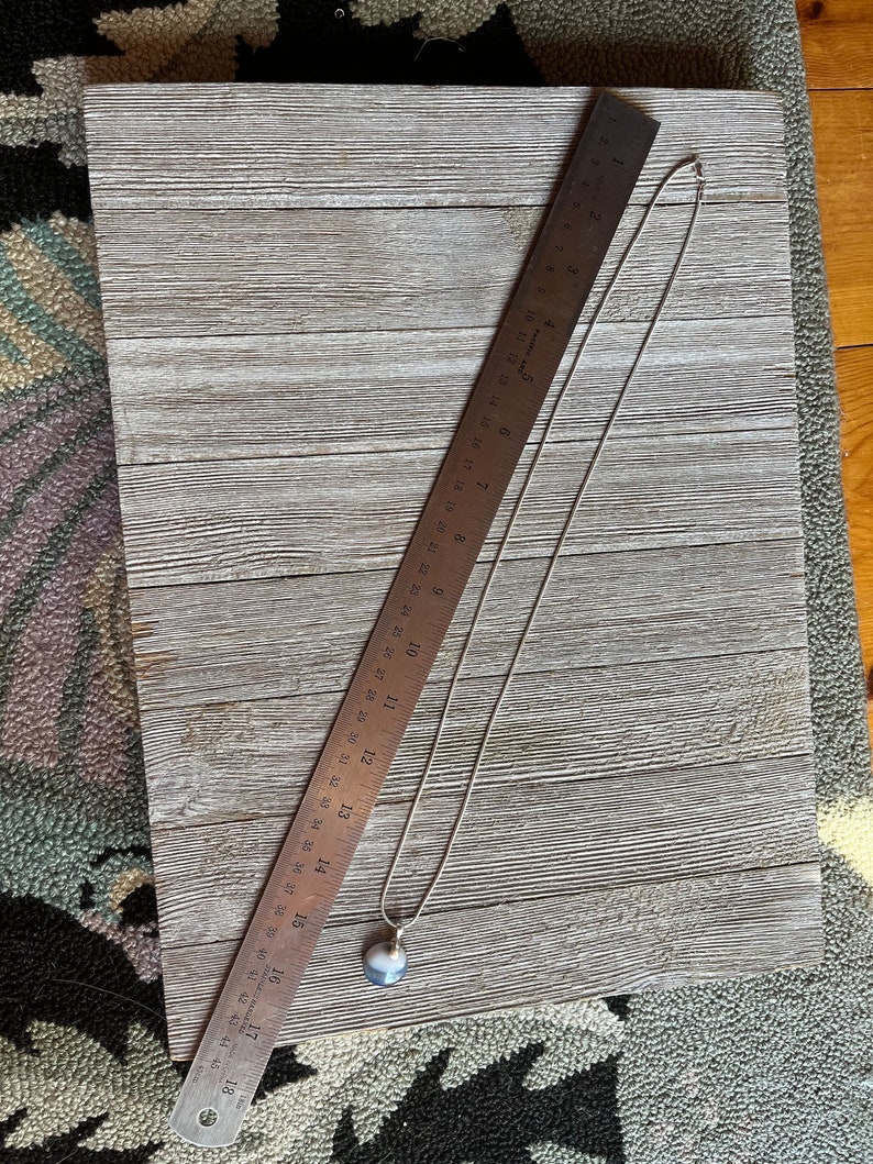 May include: A silver chain necklace with a round, gray and white agate pendant. The necklace is laying on a wooden surface with a ruler next to it.