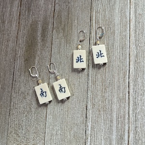 May include: A pair of earrings featuring white ceramic tiles with black Chinese characters. The tiles are rectangular and have a slightly textured surface. The earrings are silver and have a hook closure.