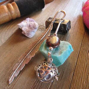 May include: A silver metal bookmark with a feather design, a brown bead, and a decorative pendant with an Om symbol. The bookmark rests on a wooden surface with a blue-green stone and other objects in the background.