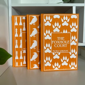 May include: Three hardcover books with orange covers. The covers feature white designs: chess pieces, ravens, and paw prints. The book on the right has the title "THE FOXHOLE COURT" and the author's name "NORA SAKAVIC".