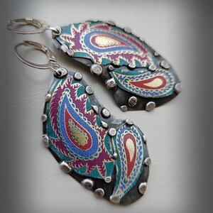 May include: A pair of dangle earrings featuring dark oxidized metal plates with silver-toned rivets along the edges. The surface displays a colorful paisley pattern in shades of magenta, teal, blue, and gold. Each earring has a silver-toned lever back.