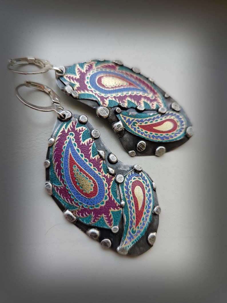 May include: A pair of dangle earrings featuring dark oxidized metal plates with silver-toned rivets along the edges. The surface displays a colorful paisley pattern in shades of magenta, teal, blue, and gold. Each earring has a silver-toned lever back.