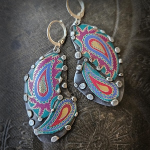 May include: A pair of dangle earrings featuring dark oxidized silver metal bases with irregular edges and small silver studs. The surface is decorated with vibrant, multi-colored paisley patterns in shades of magenta, teal, blue, and gold.