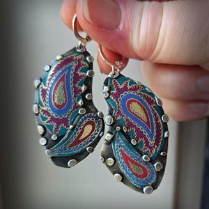 May include: A pair of dangle earrings featuring irregular, dark metal shapes with a textured surface. Each earring displays a vibrant, multi-colored paisley pattern in shades of magenta, teal, blue, and gold, accented by small, raised silver-toned metal studs along the edges.