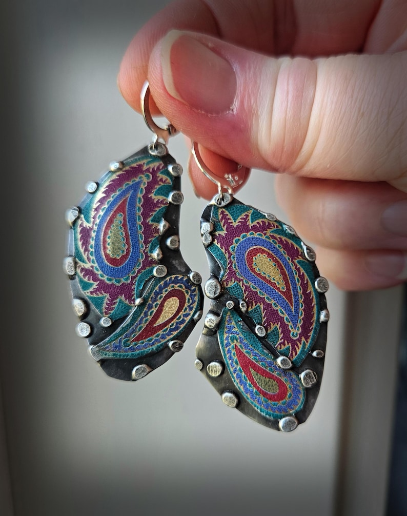 May include: A pair of dangle earrings featuring irregular, dark metal shapes with a textured surface. Each earring displays a vibrant, multi-colored paisley pattern in shades of magenta, teal, blue, and gold, accented by small, raised silver-toned metal studs along the edges.