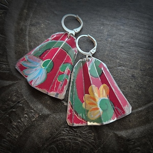 Może przedstawiać: Para ręcznie robionych kolczyków z kwiatowym wzorem. Kolczyki mają czerwone tło z zielonymi i żółtymi malowanymi kwiatami. Kolczyki mają srebrne, zawiasowe haczyki na uszy.