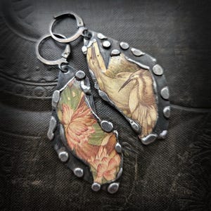 Może przedstawiać: Srebrne kolczyki w kształcie łezki. Każdy kolczyk ma nadruk w stylu vintage z ptakiem i kwiatowym wzorem, obramowany teksturowaną metalową obwódką z małymi, okrągłymi akcentami. Kolczyki mają zapięcie typu zatrzask.