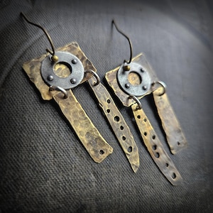 Może przedstawiać: Para ręcznie robionych kolczyków o unikalnym, industrialnym designie. Każdy kolczyk ma młotkowany mosiężny prostokąt, srebrny okrągły akcent i mosiężny wisiorek z wywierconymi otworami. Kolczyki wiszą na prostym drucianym haczyku.