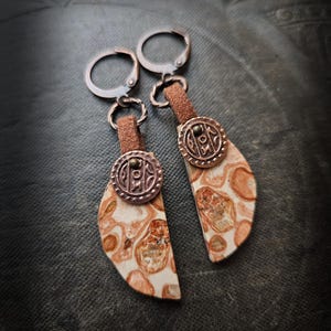 Może przedstawiać: Para ręcznie robionych kolczyków z kamieniem w kształcie półksiężyca z kremowo-brązowym wzorem. Kolczyki mają miedziane metalowe akcenty, w tym dekoracyjny okrągły element i zapięcie zatrzaskowe. Są połączone z brązowym skórzanym paskiem.