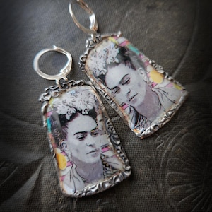 Może przedstawiać: Para srebrnych kolczyków z portretem Fridy Kahlo. Prostokątne wisiorki mają ozdobną srebrną ramkę i zapięcie zatrzaskowe. Obraz Fridy Kahlo ma kolorowe akcenty.