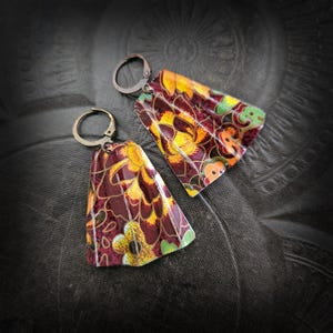 Może przedstawiać: Ręcznie robione kolczyki z kwiatowym wzorem. Kolczyki mają ciemnobordowe tło z żółtymi, pomarańczowymi i zielonymi wzorami kwiatowymi. Mają prostokątny kształt z zakrzywioną górą i brązowymi haczykami na uszy. Kolczyki mają około 3 cm długości.